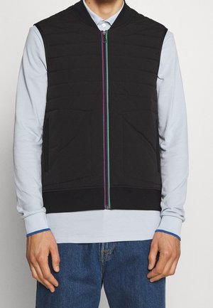 Homme portant une chemise bleu clair, un gilet noir sans manches avec fermeture éclair colorée, et un jean bleu, debout devant un fond uni clair.