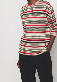 Pletený pruhovaný top s horizontálními pruhy v červené, zelené, béžové a černé barvě s metalickými akcenty; kulatý výstřih; rukávy o délce tří čtvrtin.