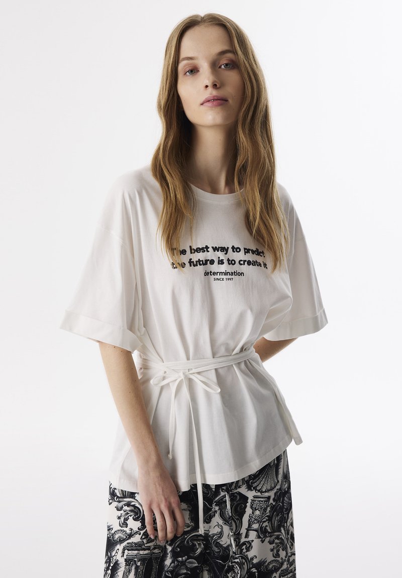 Weißes, übergroßes T-Shirt mit umgeschlagenen Ärmeln, auf der Vorderseite mit schwarzem Text. Am Taillengürtel mit einem weißen Stoffgürtel gebunden, kombiniert mit einem gemusterten Rock.