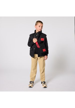 Gilet imbottito nero con loghi Hugo rossi, indossato sopra una felpa nera. Pantaloni beige e sneakers multicolore completano l'outfit.