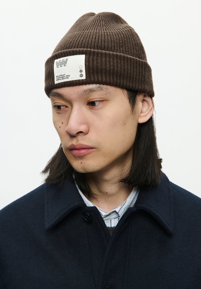 Ruda su raišteliais skrybėlė su baltu etiketu, ant kurio pavaizduota prekės ženklo logotipas ir detalės, dėvima per tamsius ilgus plaukus. Mėlynas paltas su apykakle matomas.