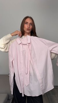 Frau mit braunen Haaren hält ein rosa gestreiftes Hemd mit Knöpfen und eine passende Krawatte an einem Kleiderbügel in einer neutralen Innenumgebung.