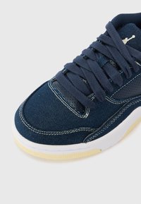 Gros plan d'une basket bleu marine avec des lacets épais, des coutures blanches, un panneau latéral perforé et une semelle blanche avec une bande de roulement beige.