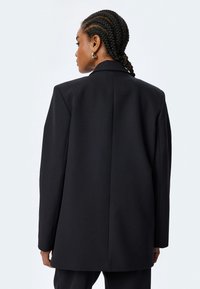 Zwarte blazer, tailored fit, gestructureerde schouders, soepele stof, enkele split aan de achterkant, minimalistisch ontwerp zonder zichtbare versieringen.
