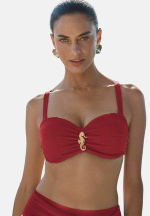 Vrouw met donker haar in een rode, gestructureerde bikini top versierd met een gouden zeepaardje, met grote oorbellen, tegen een effen lichte achtergrond.