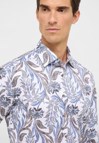Witte shirt met ingewikkelde blauwe en bruine bloemenpatronen, een kraag, en bruine knopen, met een gladde stoftextuur.