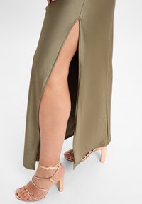 Olijfgroene satijnen maxi-jurk met een zijsplit, gecombineerd met naakte, gestrikte pumps, die een gladde textuur en een strak ontwerp benadrukken.