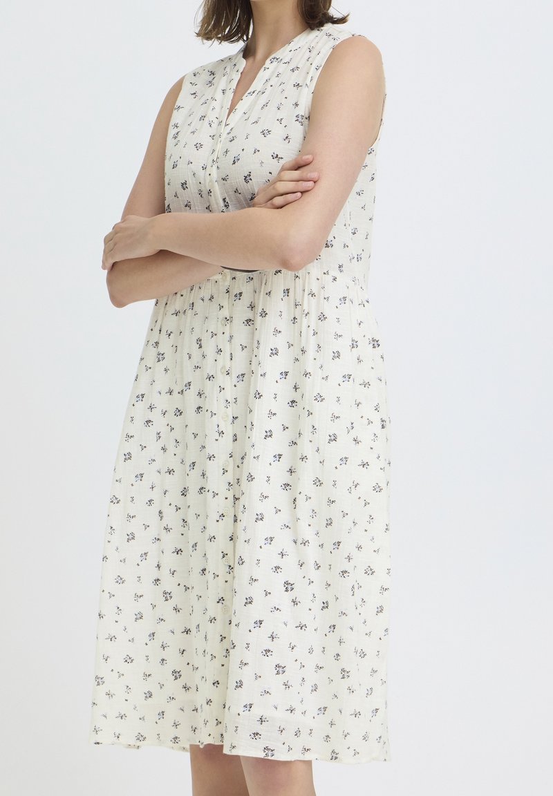 Femme portant une robe blanche sans manches avec un petit motif floral noir, debout les bras croisés devant un fond uni.