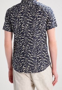 Camicia a maniche corte realizzata in tessuto scuro con un motivo a foglie color crema, dotata di colletto e orlo curvo sul fondo.