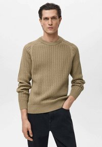 Beige gebreide trui met een ribbelstructuur, ronde halslijn en lange mouwen. Draag met zwarte broek, wat een getailleerde silhouet benadrukt.