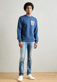 Marineblaues Sweatshirt mit pinkem Grafikdruck, Rundhalsausschnitt, gerippte Bündchen, kombiniert mit hellblauen Jeans und weißen Sneakers auf einem Holzboden.