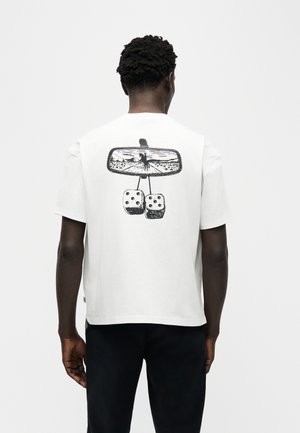 Mees, kes kannab valget t-särki, millel on must graafika auto tahavaatepeeglist, mis näitab ratsanikku hobusel ning taga ripuvad karvased täringud.