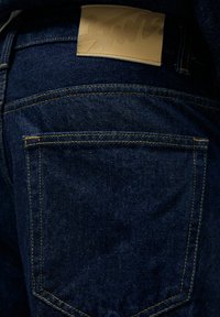 Mørkeblå denim jeans med en glat tekstur, orange syninger og et brunt lædermærke i taljen med subtil branding.