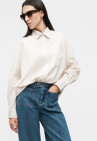 Γυναίκα που φοράει υπερμεγέθη ανοιχτόχρωμο ριγέ πουκάμισο, ψηλόμεσα μπλε τζιν με εμπρός τσέπες και σκούρα γυαλιά ηλίου, χέρι στην τσέπη.