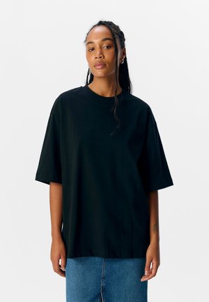 Jeune femme aux cheveux tressés portant un T-shirt noir ample et un jean bleu, debout devant un fond blanc uni.