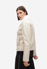 Giacca in pelle beige chiaro con colletto in pelliccia sintetica bianca, dotata di zip dorate e tasche laterali; texture liscia e design corto.