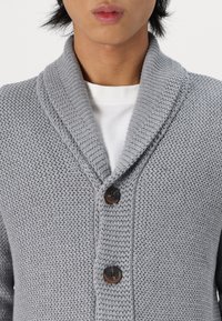 Hombre con un cárdigan de punto gris con cuello de chal y dos botones sobre una camisa de cuello redondo blanca.