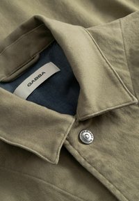 Lys beige jakke med et struktureret stof. Har en krave, der kan foldes ned, blå foer og en sølvfarvet metal knap med logodetalje.