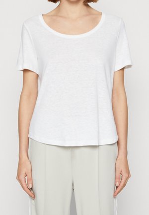 Mujer con una camiseta blanca sencilla de manga corta y cuello redondo combinada con pantalones ajustados de color gris claro, de pie frente a un fondo liso.