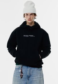 Svart hoodie med en ficka fram, prydd med vit broderad text. Matcher med ljusblå denimjeans och en vit ribbad mössa.