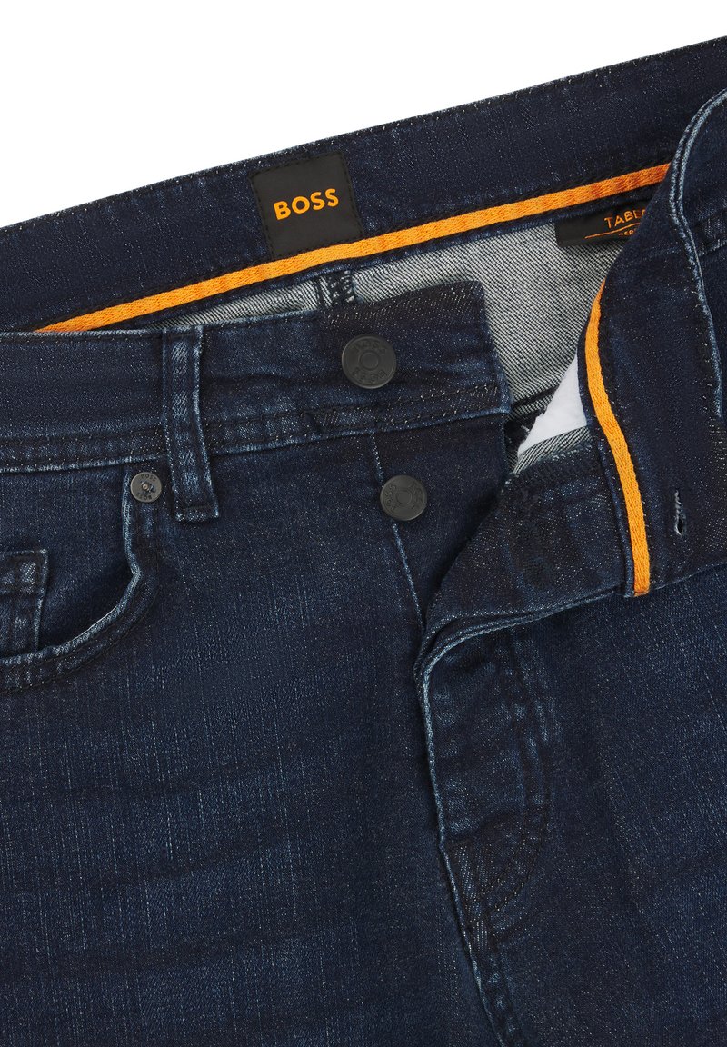 Dunkelblaue Jeans aus Denim mit glatter Textur, doppelter Knopfverschluss und kontrastierendem orangefarbenem Nähen im Innenbund. Markenlabel sichtbar.