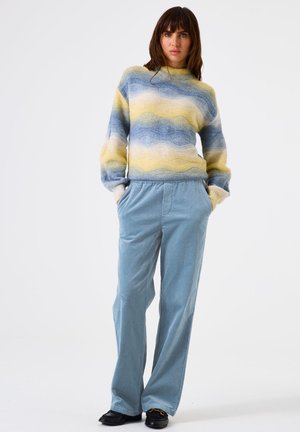 Woman standing with hands in pockets wearing a wavy striped sweater in blue and yellow, light blue pants, and black loafers against white background.