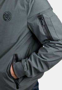 Chaqueta gris con una textura suave, que presenta un parche con el logo, un bolsillo con cremallera en la manga y bolsillos laterales con detalles en negro.