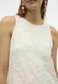 Vit, ärmlös topp med texturerade, blomapplikation detaljer. Mjukt, lättviktigt tyg med en något skrynklig yta.