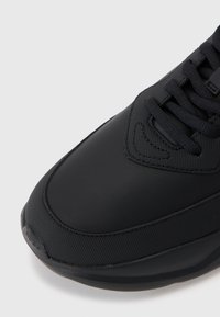 Svart sneaker med en glatt, matte overflate og teksturerte detaljer. Designet inkluderer lisser og en polstret såle.