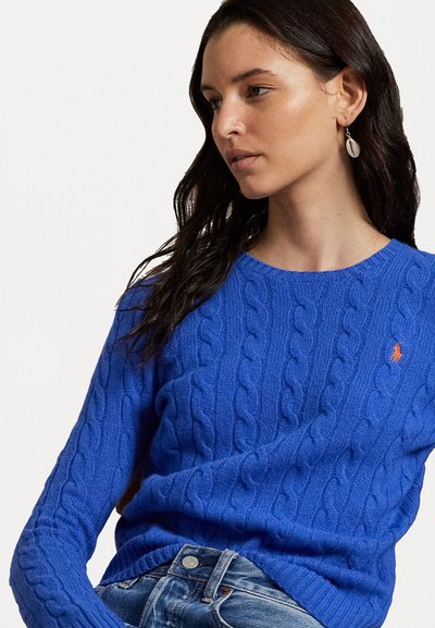 Blå kabelstrikket genser med ribbestrikket hals og nederkant, prydet med en brodert oransje logo på brystet, sammen med denimjeans.