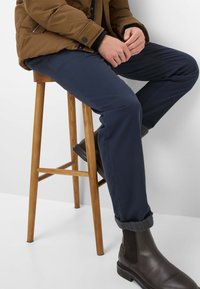 Pantalons slim-fit navy avec des manches retroussées, accompagnés de bottines en cuir marron. Une doudoune marron est portée sur le haut du corps.