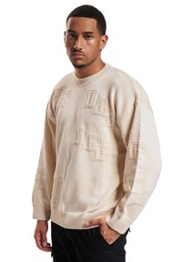 Maglione in maglia beige con un design testurizzato caratterizzato da lettere in rilievo ripetute. Le maniche a costine e l'orlo completano il look.