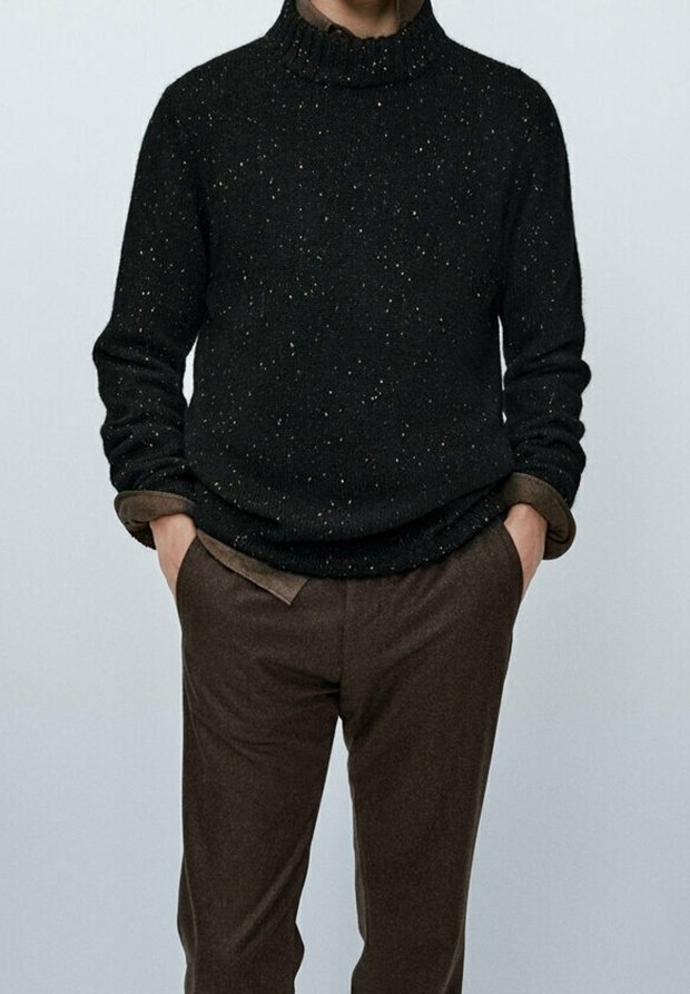 Personne portant un pull à col roulé tacheté foncé sur une chemise marron avec un pantalon marron, les mains dans les poches, debout devant un fond uni.
