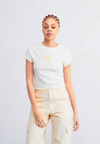 Witte geribbelde crop top met geschulpte randen, versierd met een kleine gele bloemenborduur. Draag het met lichtbeige cargobroek.
