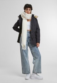 Mornarska zimska parka s kapuco in umetnim krznom, v kombinaciji s smetanasto pleteno šaljico, belim kratkim topom, širokimi svetlo modrimi kavbojkami in belimi športnimi copati.