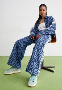 Blåmönstrad denimjacka och vida jeans med vita ränder, ihop med ljusblå sneakers, sittande på en brun stol mot en enfärgad bakgrund.