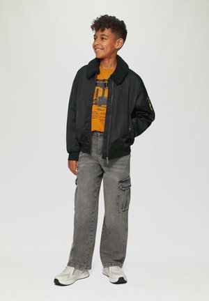 Schwarze Bomberjacke mit einem Schaffellkragen, orangefarbenes Grafik-T-Shirt, graue Cargo-Hosen und helle Sneaker. Einfaches Design und lässige Passform.