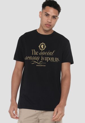 Young man wearing a black t-shirt with gold text: "The social season is upon us. Bridgerton" and a Victorian-style silhouette above.