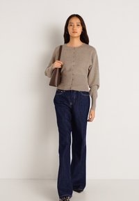 Καφέ πλεκτό πουλόβερ με κουμπιά μπροστά και ριμπ σχέδιο στο στρίφωμα, συνδυασμένο με σκούρα μπλε καμπάνα τζιν. Μοντέλο κρατάει μια καφέ τσάντα.