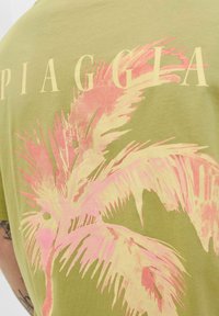 Olivenfarvet t-shirt med et stort pink palmebladtryk og ordet "PIAGGIA" i lyse farver på ryggen. Blødt bomuldsstof.
