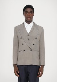 Szürke, kétsoros zakó fekete gombokkal, hajtókás gallérral és két oldalzsebbel. Fehér galléros ing fölött és sötétkék csíkos nadrágban viselve.