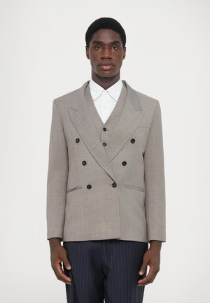 Grau doppelt gepunkteter Blazer mit schwarzen Knöpfen, mit Reverskragen und seitlichen Taschen. Getragen über einem weißen Hemd mit Kragen und gestreiften Hosen.