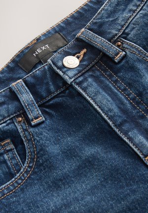 Donkere blauwe spijkerbroek met contraststiksels, knoopsluiting en een geweven label. Gladde textuur met lichte vervaging op sommige plekken.