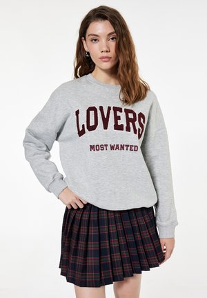 Grijze sweatshirt met "LOVERS MOST WANTED" in bordeauxrode letters, gecombineerd met een zwarte geplooide rok met een ruitpatroon en rode accenten.