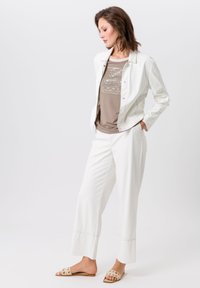 Witte jas met zilverkleurige hardware, beige graphic t-shirt en witte brede broek. Tan sandalen met ronde uitsparingen. Soepele texturen overal.