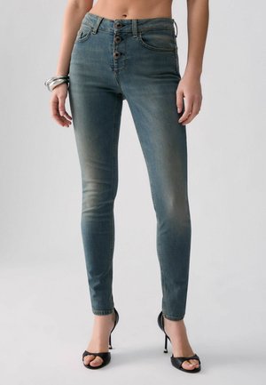 Femme portant un jean skinny bleu délavé taille haute avec trois boutons, des talons ouverts noirs et des bracelets argentés, debout devant un fond uni.