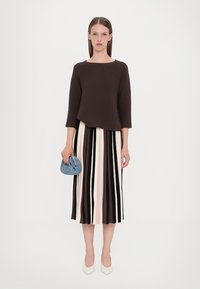 Pull en maille marron à manches trois-quarts, associé à une jupe midi rayée en crème, marron et rose pâle. Tenant un sac tissé bleu.