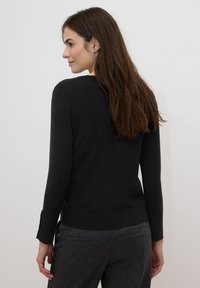 Pull noir à manches longues avec encolure ronde et fentes latérales, fabriqué dans un matériau doux, associé à un pantalon gris taillé sur mesure.