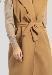 Gilet long couleur camel en tissu doux. Il présente un large col, une coupe oversize et une ceinture à nouer à la taille. Des poches visibles sont incluses.