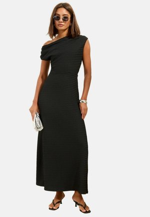 Žena má na sebe čierne textúrované maxi šaty so skoseným jedným ramienkom, čierne sandále na opätku, slnečné okuliare, strieborný náramok a drží striebornú kabelku.